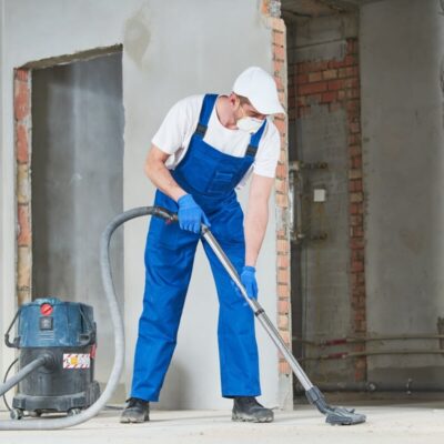 Pracownik w kombinezonie wykonuje sprzątanie po budowie za pomocą odkurzacza przemysłowego.