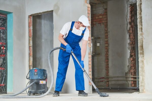 Pracownik w kombinezonie wykonuje sprzątanie po budowie za pomocą odkurzacza przemysłowego.