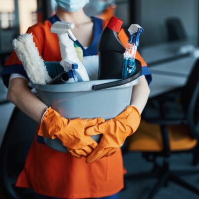 Osoba w stroju roboczym i rękawiczkach trzyma wiadro z przyborami, symbolizując usługi sprzątające dla firm.